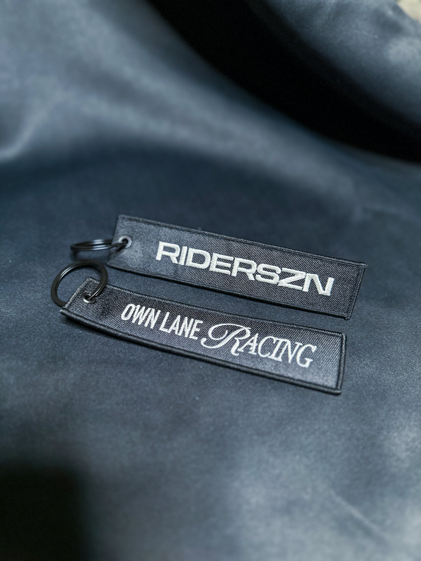 Two black jet tags with white text, one tagged 'RIDERSZN' and the other 'OWN LANE RACING', placed on a textured dark background.