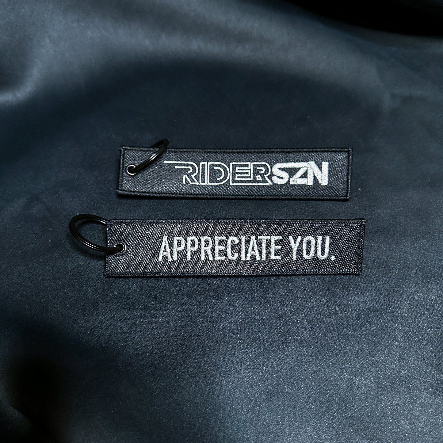 Two black embroidered jet tags attached to a dark fabric; one tag reads 'RIDER SZN' and the other 'APPRECIATE YOU'.