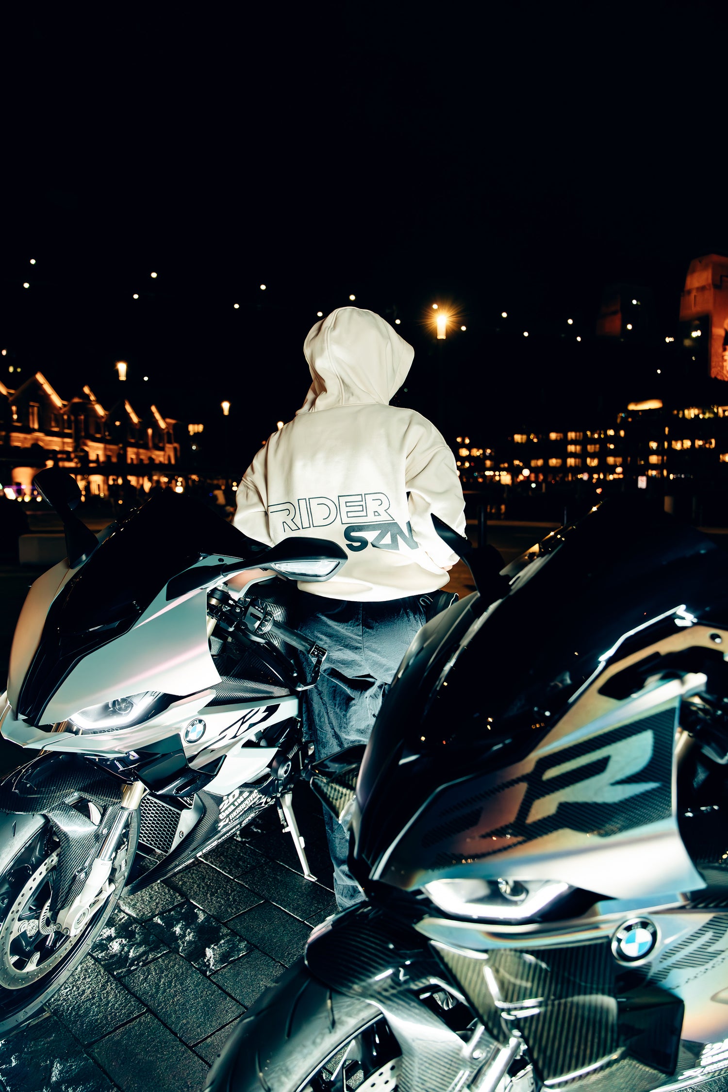 back of a person wearing a stone "Riderszn" armoured hoodie standing between two motorcycles at night