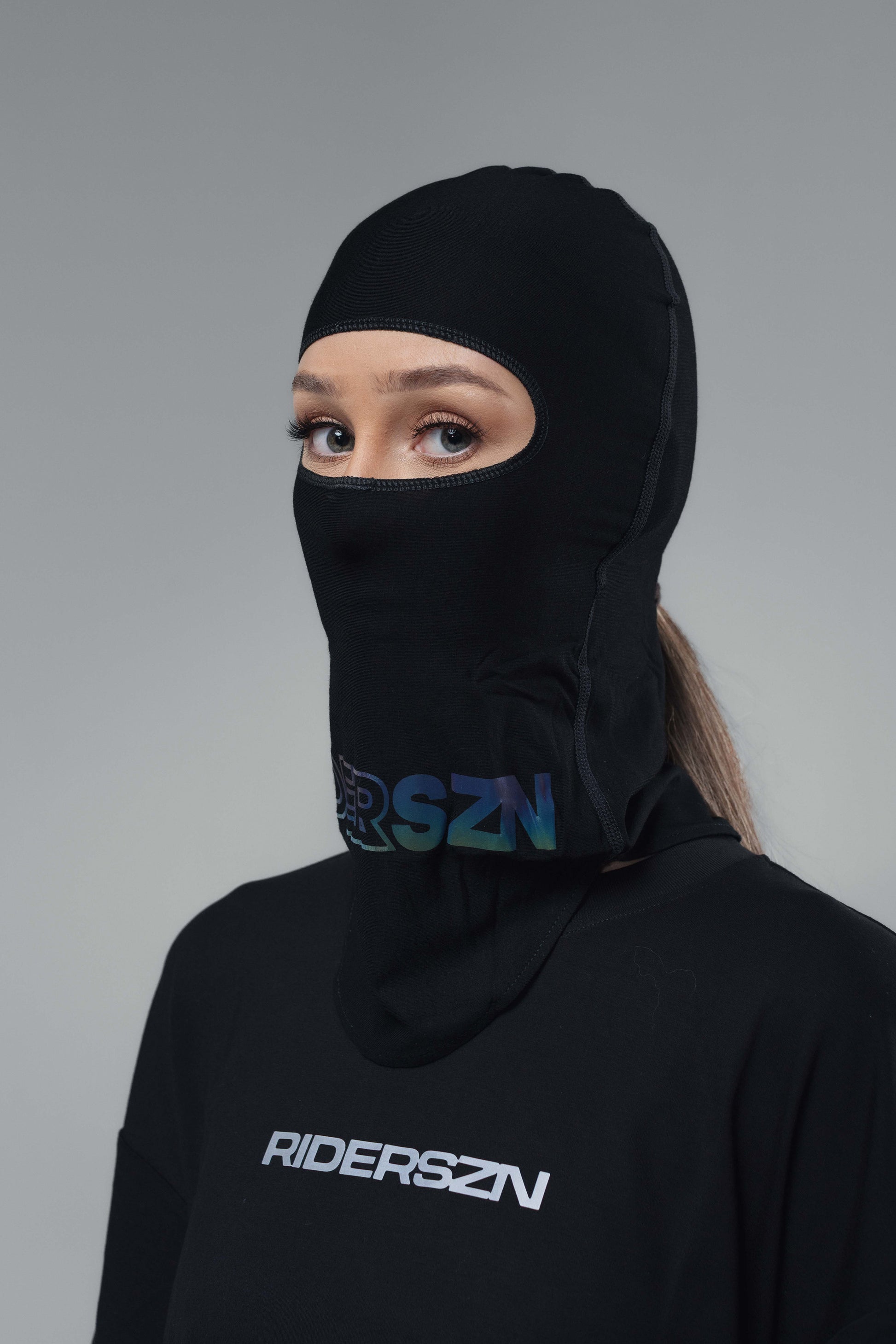 Person wearing a black soft seam balaclava with holographic 'RiderSZN' branding front view and a ponytail